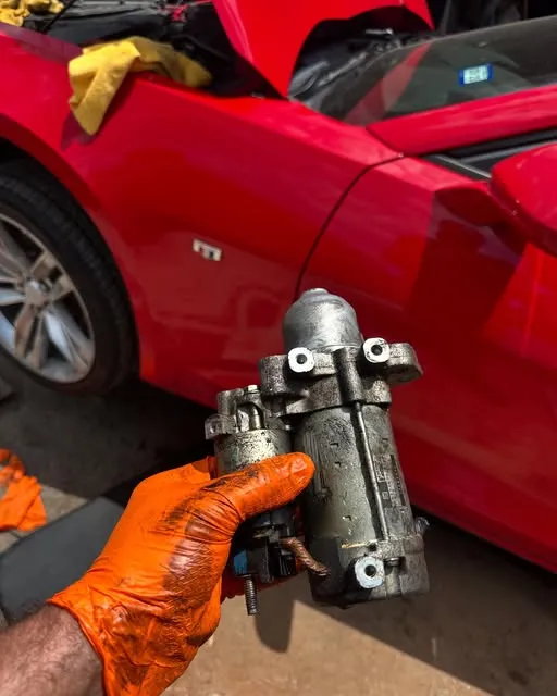 Gloved hand holding a removed starter motor beside a red car